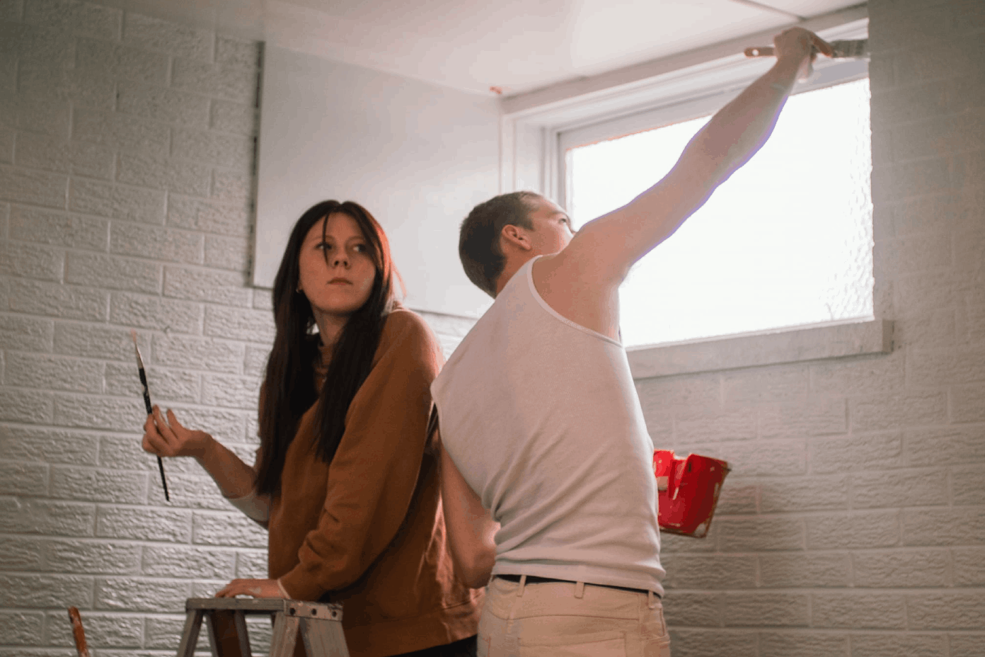 A woman and man standing on ladders painting a brick room.